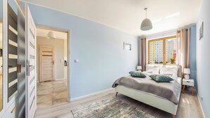 A bedroom with a bed, a window with curtains, a bedside table, and a door leading to another room. - Sun & Snow Kwartał Róży Wiatrów (Swinoujscie)