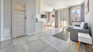 A modern living room with a grey sofa, a white side table, and a dining area with a white table and chairs. - Sun & Snow Kwartał Róży Wiatrów (Swinoujscie)