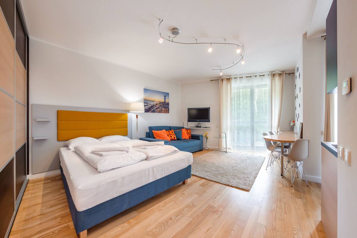 A modern hotel room with a bed, a sofa, a dining table, and a TV.