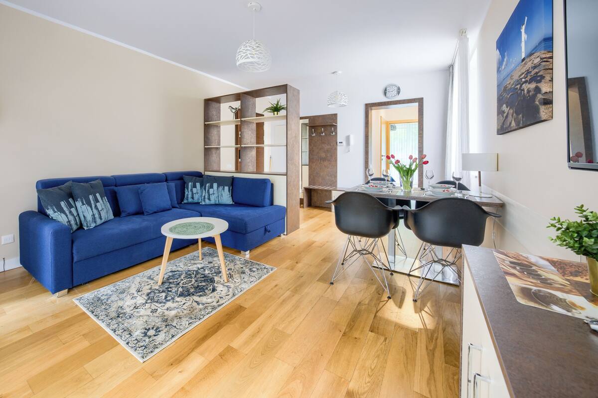 A modern living room with a blue sofa, a small round table, and a dining area with two black chairs.
