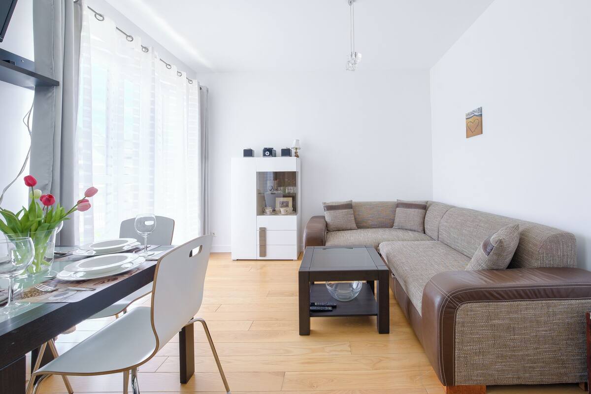 A modern living room with a brown leather sofa, a glass coffee table, and a dining area with a white table set for two.