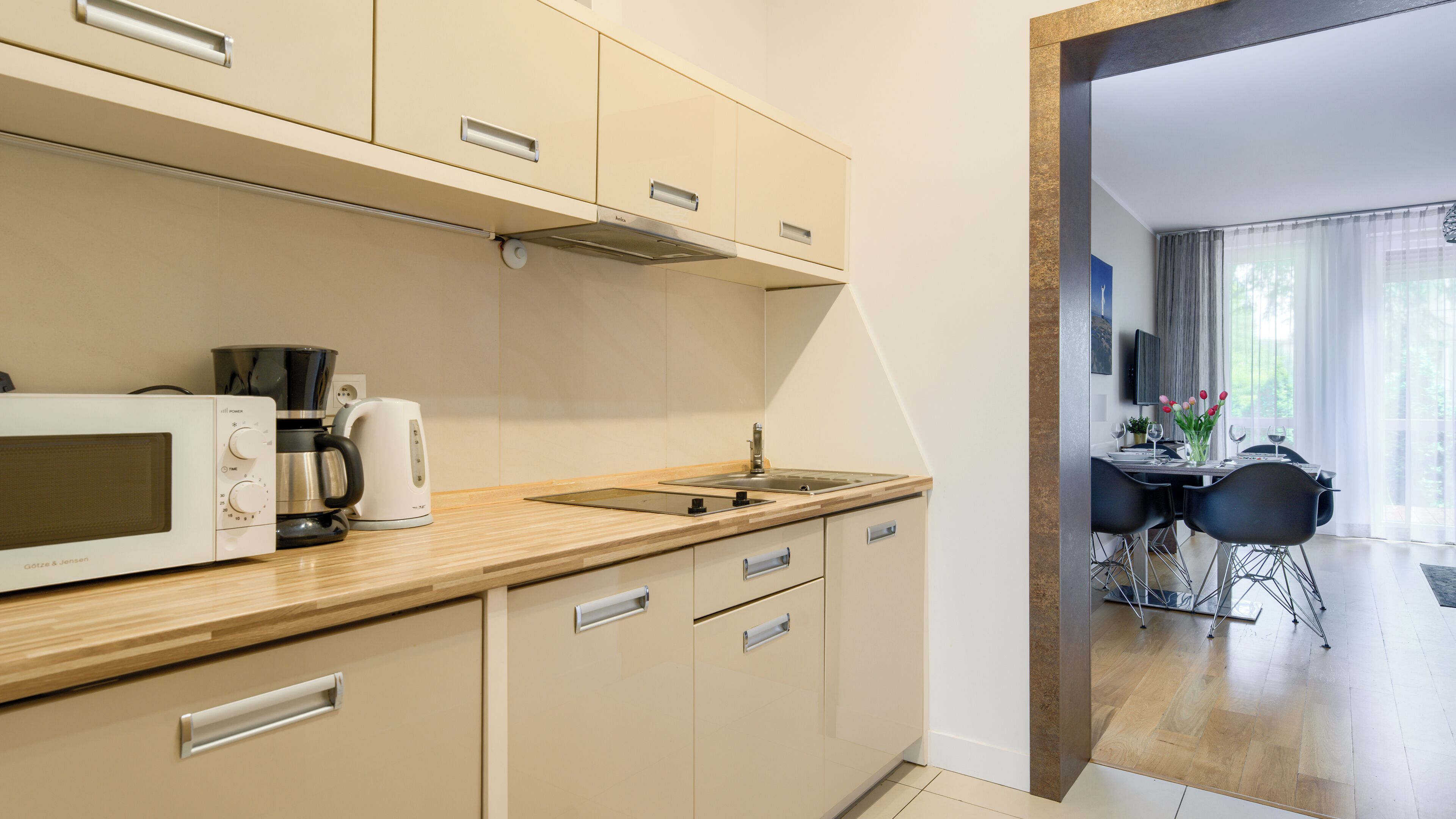 A modern kitchen with a microwave, coffee maker, and kettle. There is a sink and a dining area visible in the background.