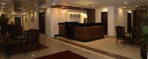 A hotel lobby with a reception desk, wooden chairs, a red carpet, and a clock on the wall.