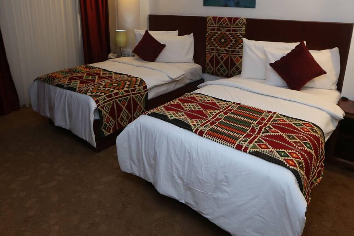 A hotel room with two beds, each with patterned bedspreads and white linens. There is a painting on the wall above the beds, a lamp on the nightstand, and a window with curtains.