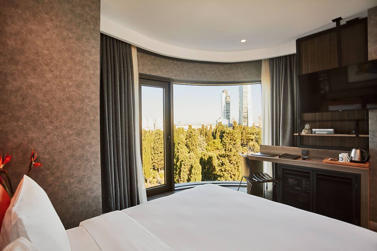 A modern hotel room with a large bed, a desk with a laptop, and a view of a cityscape through large windows.