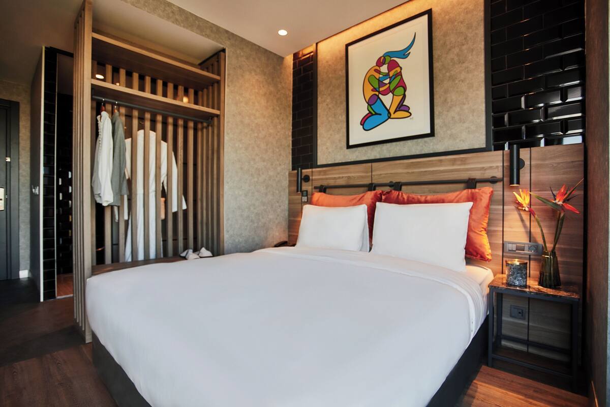 A modern hotel room with a large bed, a wooden wardrobe, a painting, and a nightstand with a vase.