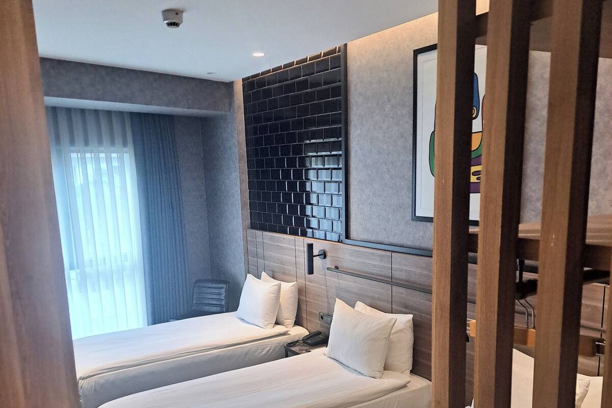 A hotel room with two beds, a large window with blinds, a black brick wall, and a framed picture on the wall.