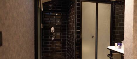 A modern bathroom with black tiled walls, a glass shower enclosure, and a white countertop with toiletries.
