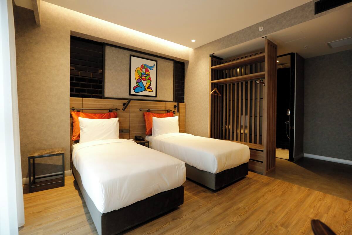 A hotel room with two beds, a wooden floor, a framed artwork above the beds, and a wardrobe with vertical slats.