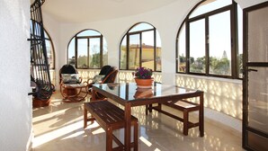 A sunlit room with a glass-top table, two wicker chairs, a bench, and large arched windows offering a view of the outdoors. - CASA CARLOS - Private Villa Detached with Pool (Cdad. Quesada)