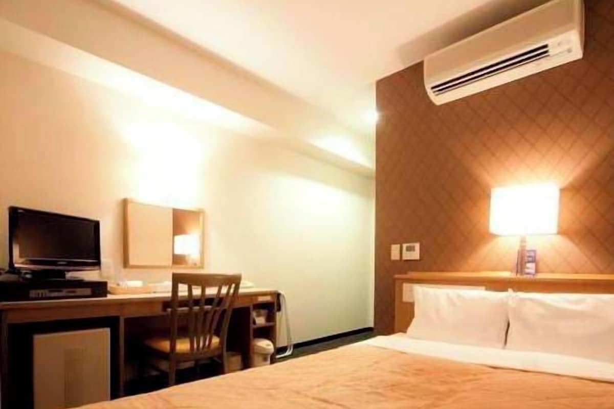 A hotel room with a bed, a desk with a chair, a television, and an air conditioning unit.