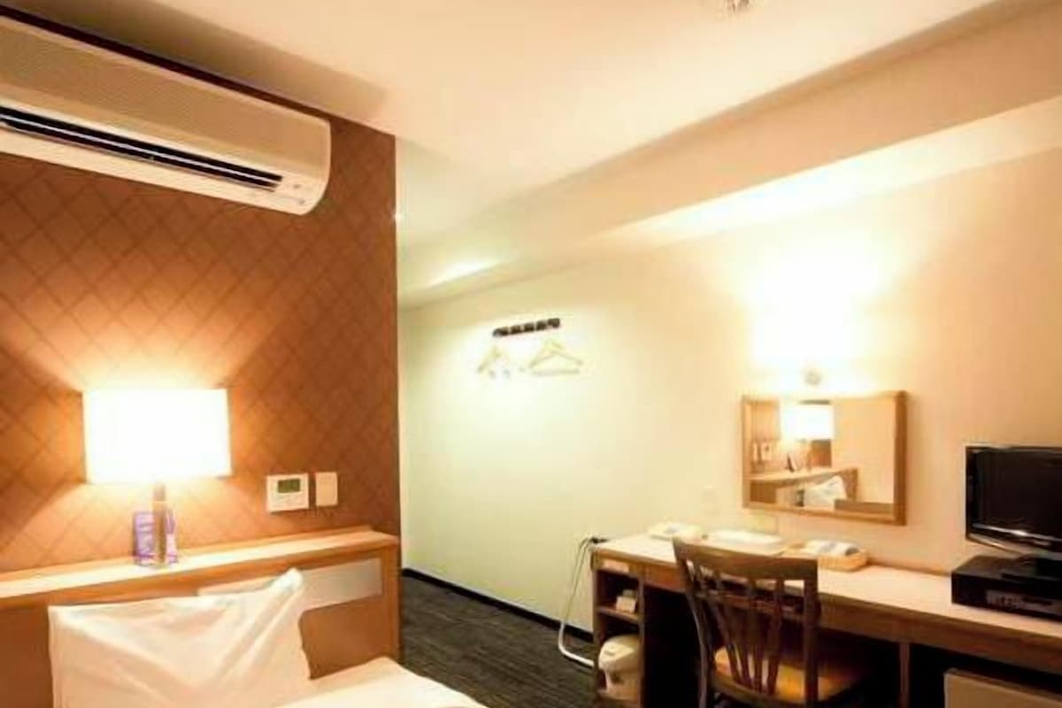 A hotel room with a bed, a desk with a chair, a television, and a wall-mounted air conditioner.