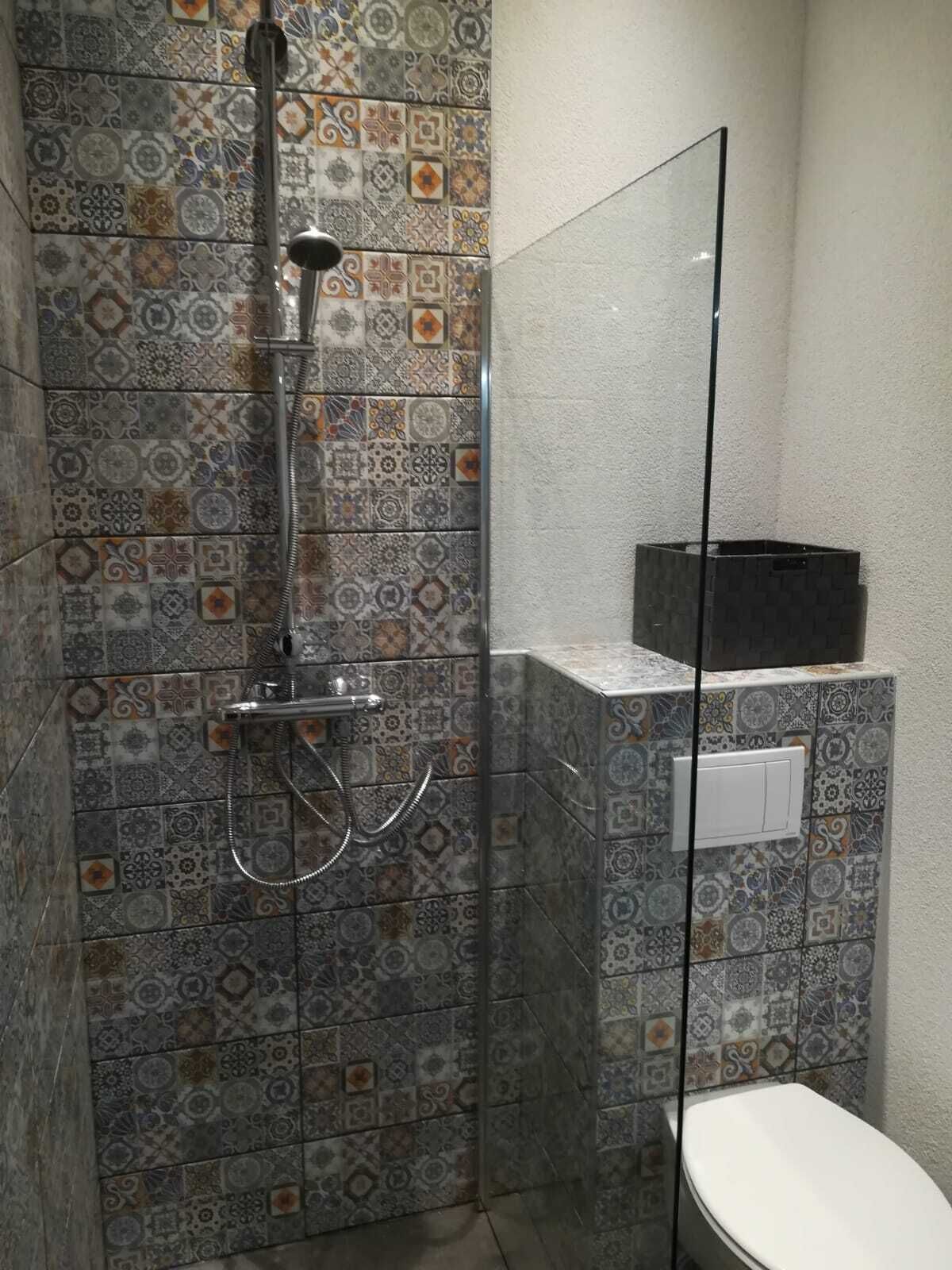 Een douche met glazen deur, betegelde wanden met een patroon en een wit toilet.