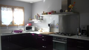 A modern kitchen with a microwave, stove, and oven. There is a window with curtains, a shelf with kitchen utensils, and a basket on the counter. - Chalet on the shore of Lake Séchemailles (Ambrugeat)