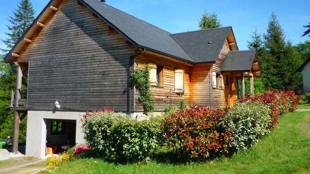 A wooden house with a sloped roof, a garage, and a well-maintained garden with colorful flowers. - Chalet on the shore of Lake Séchemailles (Ambrugeat)