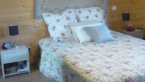 A bedroom with a floral bedspread, a nightstand with a lamp, and a tapestry on the wall. - Chalet on the shore of Lake Séchemailles (Ambrugeat)