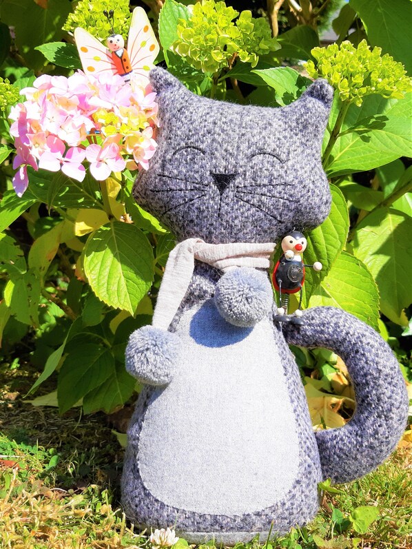 Una figura de gato tejida con nariz roja y una pequeña figura de persona sobre su lomo, ubicada frente a una planta con flores rosas y hojas verdes.
