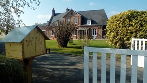 A wooden birdhouse with two bird illustrations, a white picket fence, and a brick house with a lawn in the foreground.