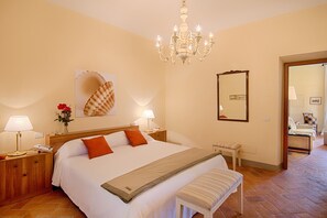 Une chambre d’hôtel avec un grand lit, des lampes de chevet, une table de nuit en bois, un banc, un lustre et un miroir au mur.
