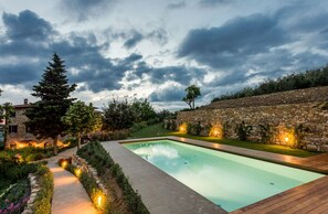 Une piscine avec des murs en pierre, une terrasse en bois et un chemin de jardin agrémenté de luminaires.