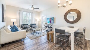 A modern living room with a white sofa, two black chairs, a glass coffee table, a dining table with chairs, a ceiling fan, and a TV on the wall. - Modern Vista Cay nr pool•Walk to OCCC close iDrive (Orlando)