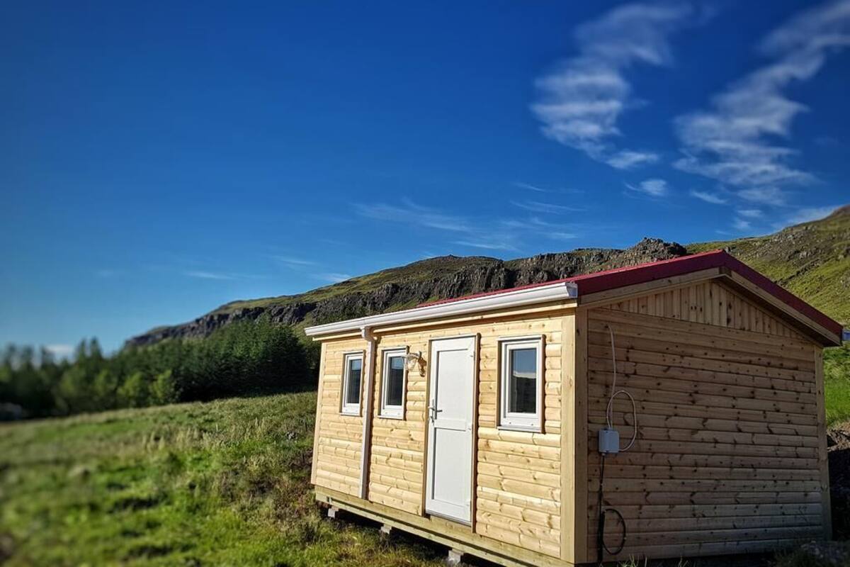 A small wooden cabin with a red roof, a door, and two windows, situated in a grassy field with a mountain in the background.