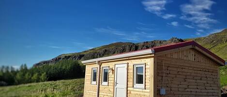 A small wooden cabin with a red roof, a door, and two windows, situated in a grassy field with a mountain in the background.