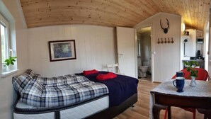 Ein gemütliches Zimmer mit einem Bett, einem kleinen Tisch mit einer Pflanze, einem Bild an der Wand und einer Küchenzeile im Hintergrund.