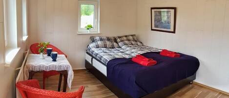 Quarto com cama, janela, quadro emoldurado, mesa pequena com cadeira e uma cadeira vermelha.