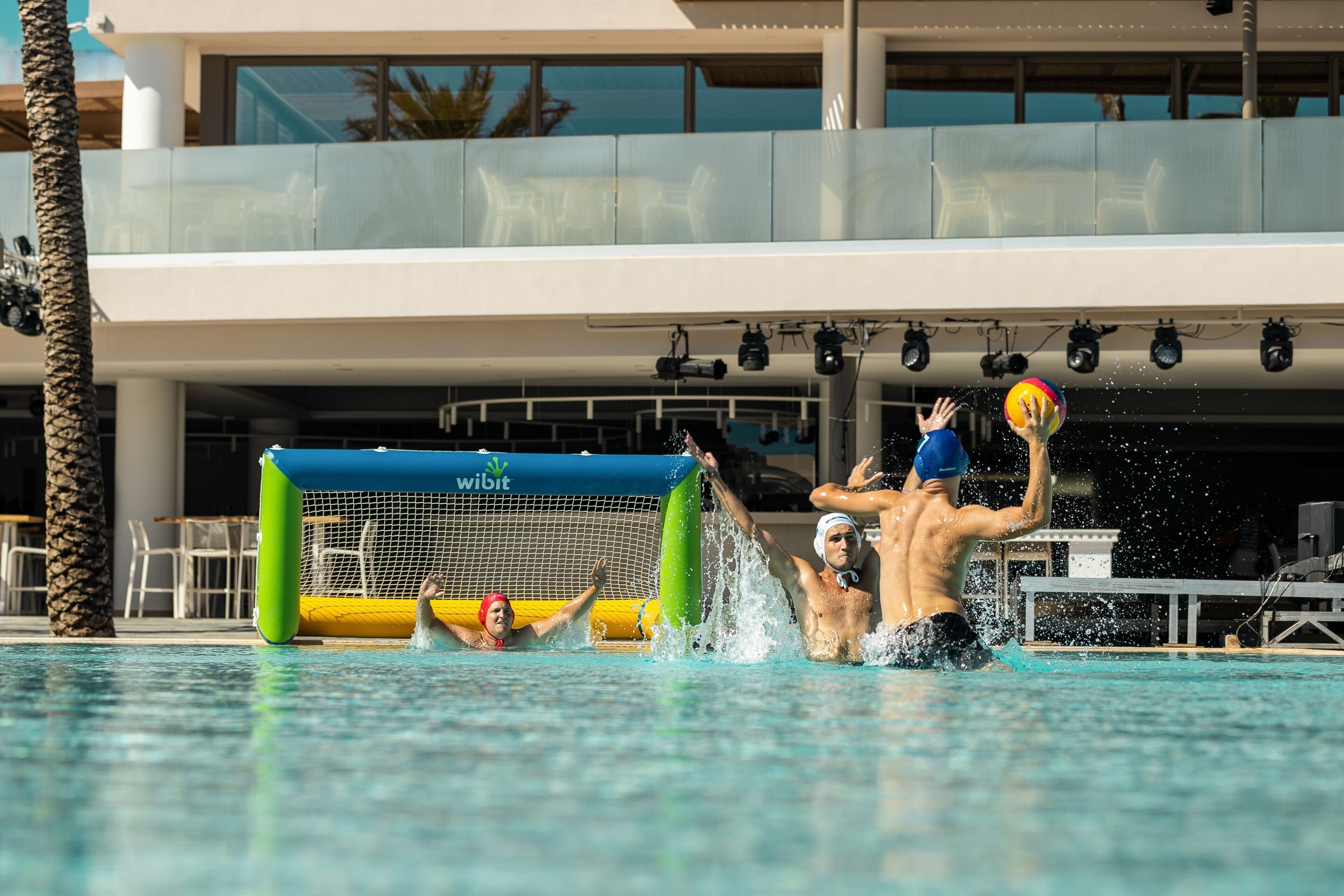 A swimming pool with a netted area for water polo, surrounded by palm trees and a modern building with large windows.