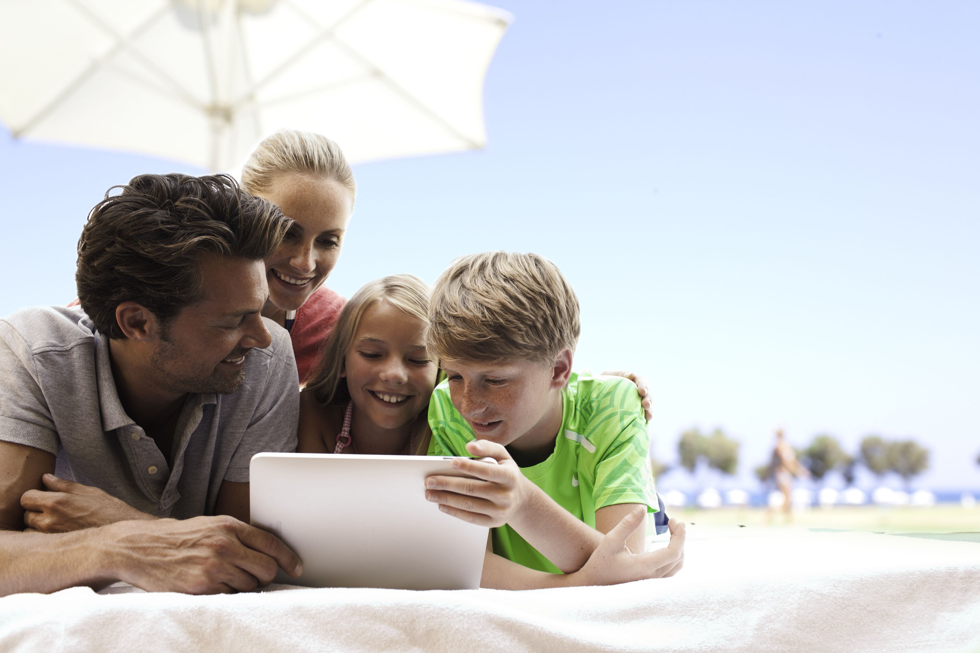 Een gezin van vier ligt op een bed met een laptop, onder een witte parasol.