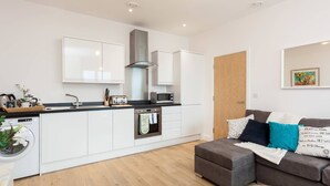 A modern kitchen with white cabinets, a built-in oven, and a microwave. There's a grey sofa with teal cushions and a framed quote on the wall. - Belfry Quarters (Redhill)