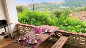 A wooden table set with pink plates and glasses, a vase of pink flowers, and a grill on a balcony overlooking a scenic view of a city and greenery. - Rustic house on montain with privileged views of Mediterranean sea (Pineda de Mar)