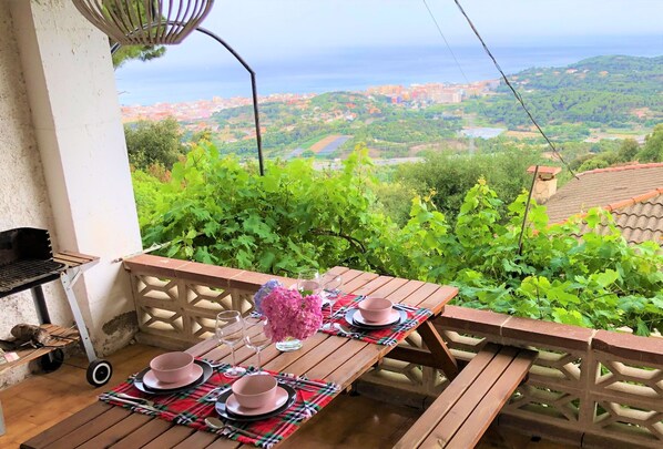 Una mesa de madera dispuesta con platos y vasos rosas, un jarrón con flores rosadas y una parrilla en un balcón con vistas panorámicas a una ciudad y vegetación.