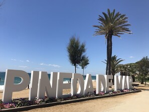 Un gran cartel con el texto "PINEDA DEW" rodeado de un parterre, con una playa y palmeras al fondo.