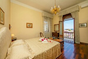 A hotel room with a bed, a desk, a chair, a chandelier, a window with curtains, and a balcony view. - Villa Barbara (Desenzano del Garda)