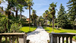 A stone pathway leading through a well-maintained garden with palm trees and a clear sky. - Villa Barbara (Desenzano del Garda)