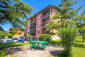 Un immeuble résidentiel à plusieurs étages, doté de balcons, d’une allée pavée et d’un espace extérieur aménagé avec des sièges.