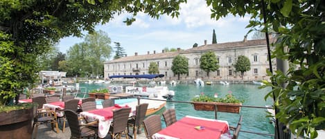 Un espace de restauration en plein air, avec des tables et des chaises, donnant sur un canal où naviguent des bateaux, et avec un bâtiment historique en arrière-plan.