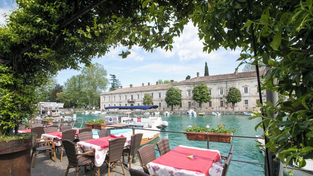 An outdoor dining area with tables and chairs, overlooking a canal with boats and a historic building in the background. - Casa dei Quadri (Peschiera del Garda)