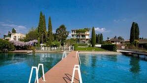 Um píer de madeira que se estende por uma piscina azul cristalina, rodeado por árvores e um edifício com varandas.