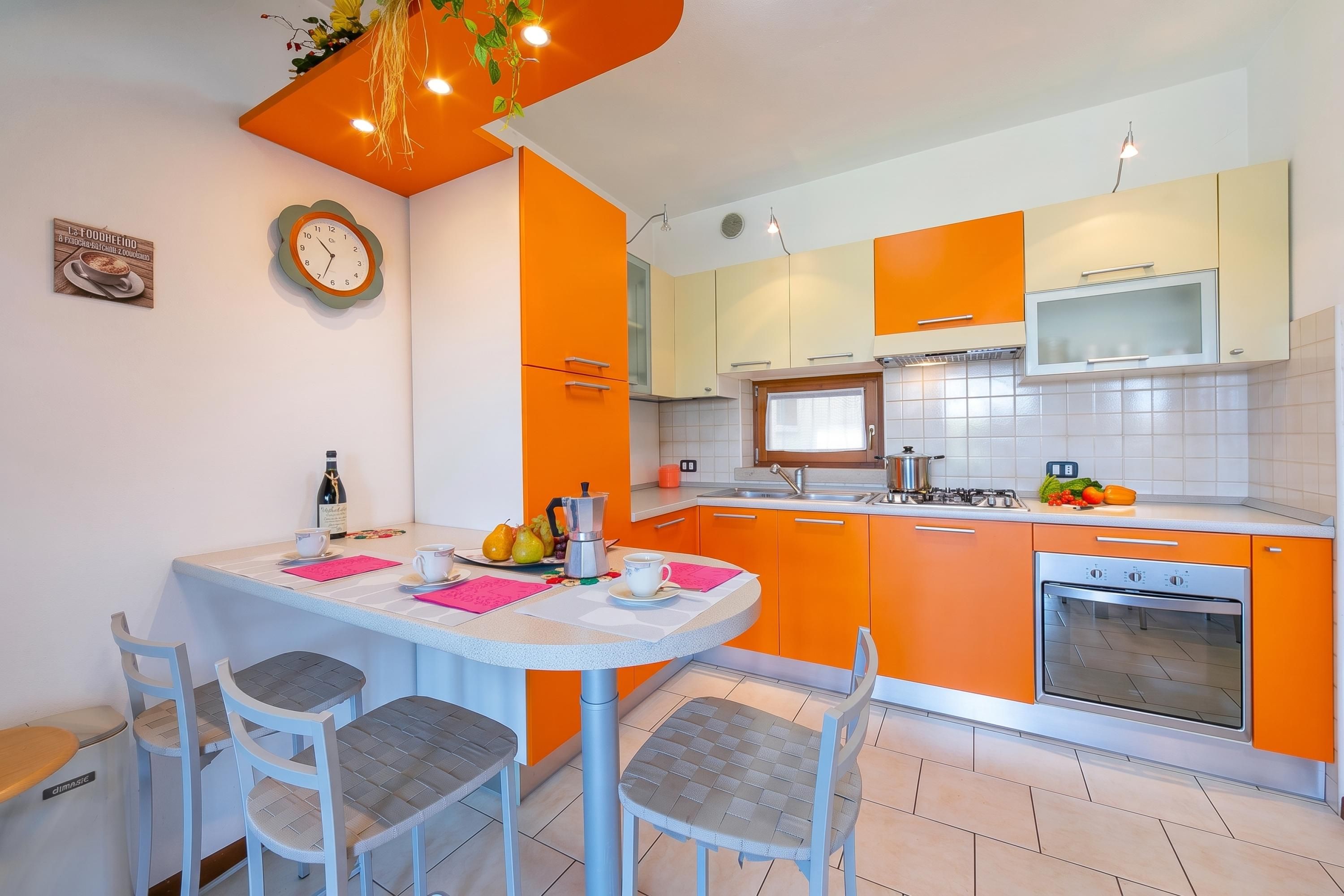 A modern kitchen with orange cabinets, a white countertop, and a built-in oven. There are two chairs and a table with a clock on the wall.