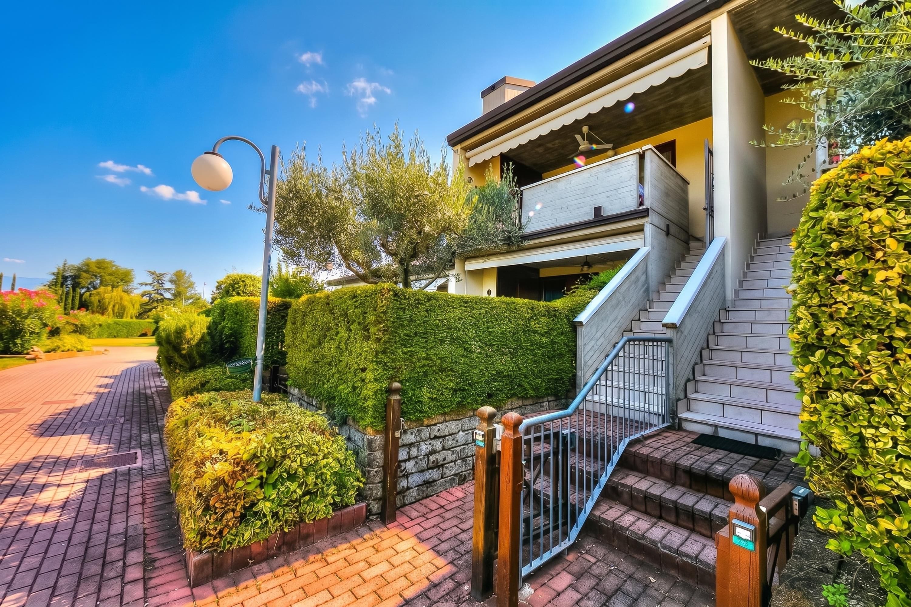 A modern building with a staircase leading to a balcony, surrounded by well-maintained hedges and a clear sky.