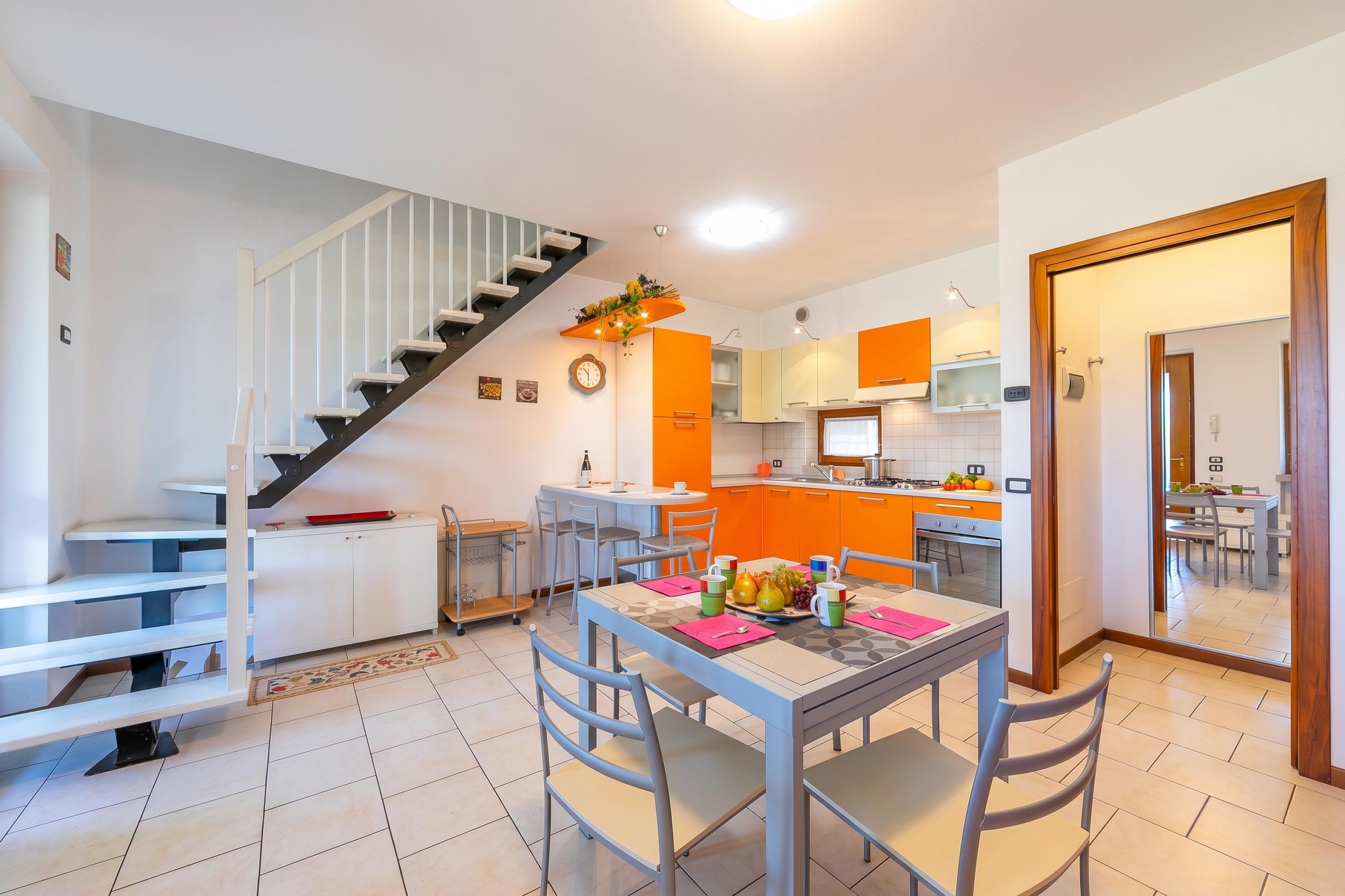 A modern kitchen with orange cabinets, a dining area with a table set for two, and a staircase with a white railing.