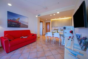 A modern living room with a red sofa, a dining area with white chairs, a kitchen with white cabinets, and a mounted TV. - Appartamento Vista Lago (Desenzano del Garda)