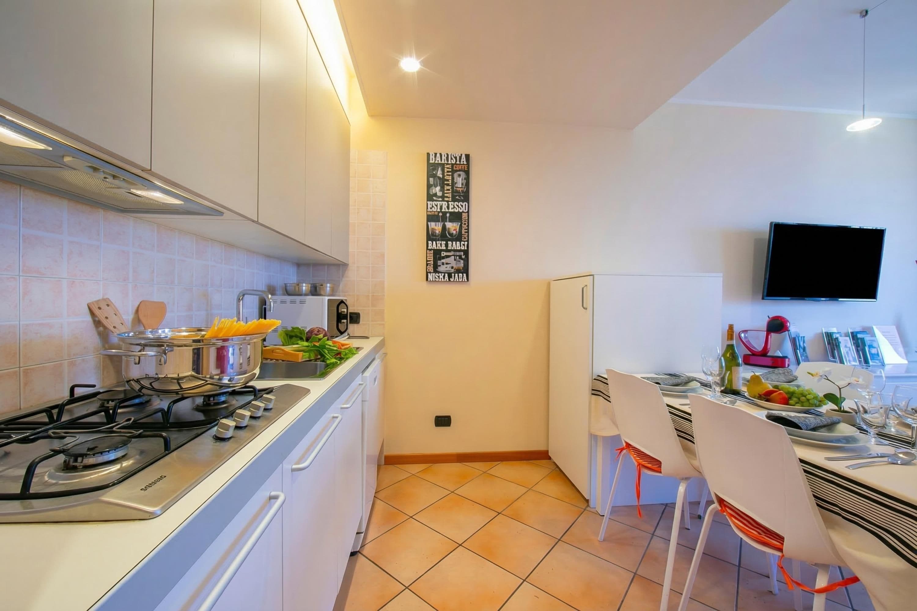 A modern kitchen with a gas stove, white cabinets, a tiled backsplash, a wall-mounted TV, and a dining area with white chairs and a table set for two.