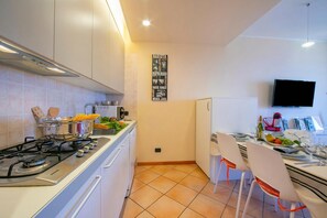 A modern kitchen with a gas stove, white cabinets, a tiled backsplash, a wall-mounted TV, and a dining area with white chairs and a table set for two.