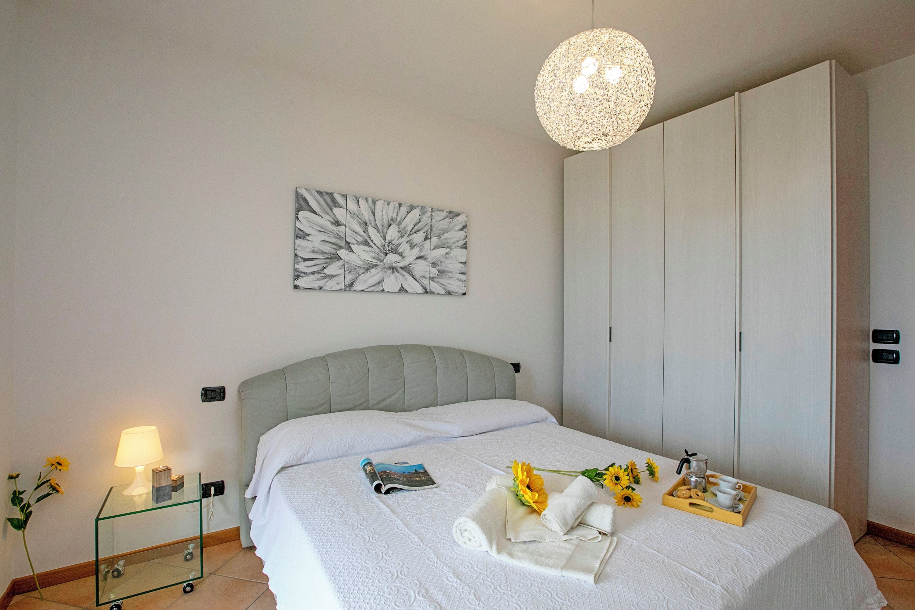 Een keurig opgemaakt bed met hoofdeinde, een nachtkastje met een lamp, een lijst met kunstwerk aan de muur, en een dienblad met handdoeken en een bloem op het bed.