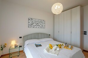 A neatly made bed with a headboard, a bedside table with a lamp, a framed artwork on the wall, and a tray with towels and a flower on the bed. - Appartamento Vista Lago (Desenzano del Garda)