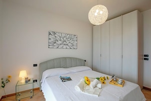 Una cama bien hecha con cabecera, una mesita de noche con lámpara, un cuadro en la pared y una bandeja con toallas y una flor sobre la cama.